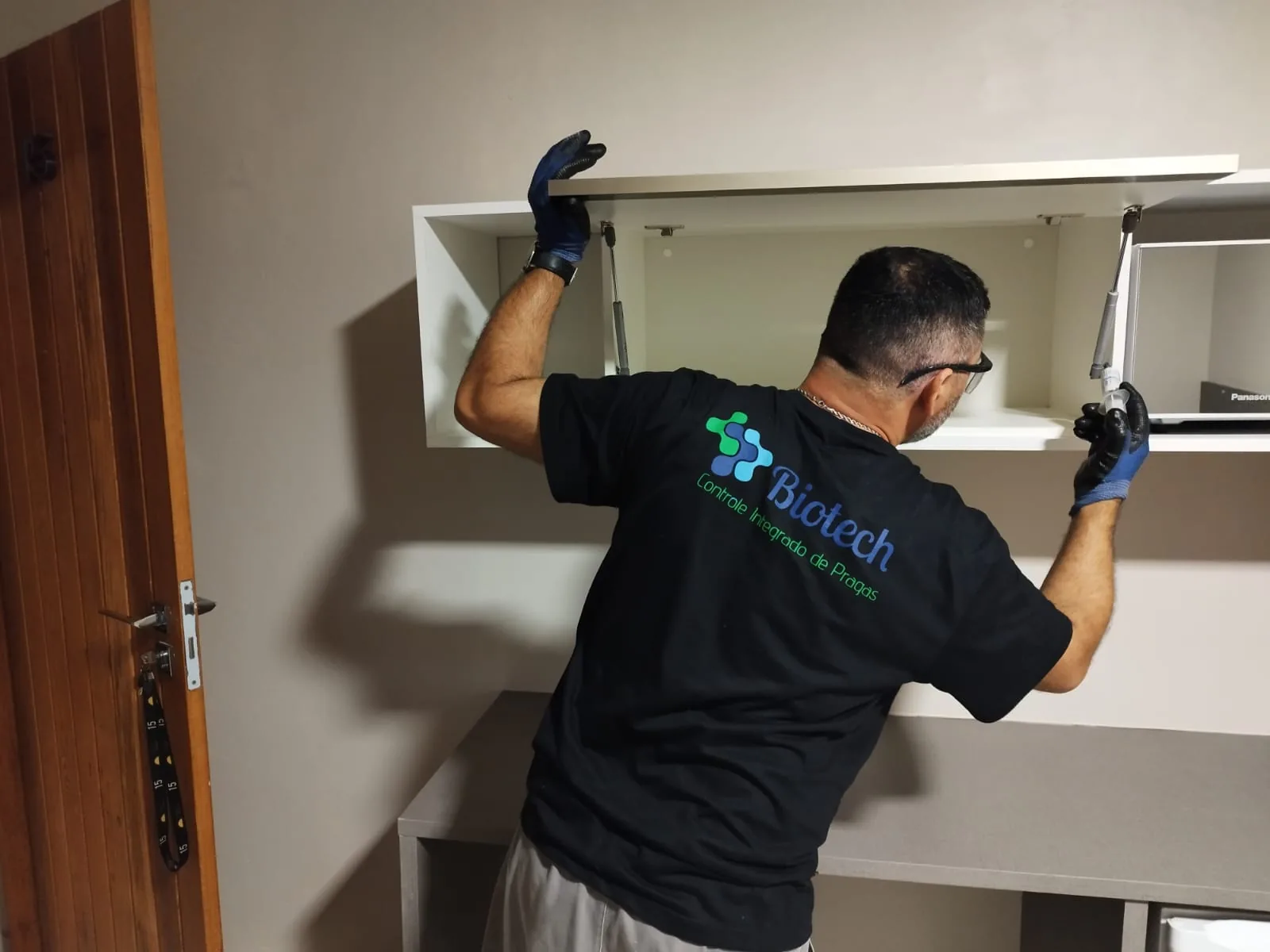 Técnico da Biotech, de camiseta da empresa e luvas, abrindo o armário de uma cozinha durante a inspeção.